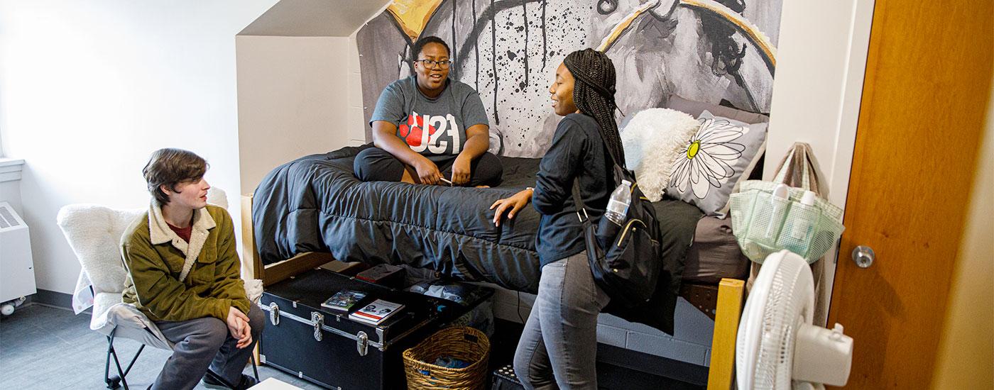 Students chat in their dorm room. One student is sitting on the bed, one is standing, and one is sitting in a chair.